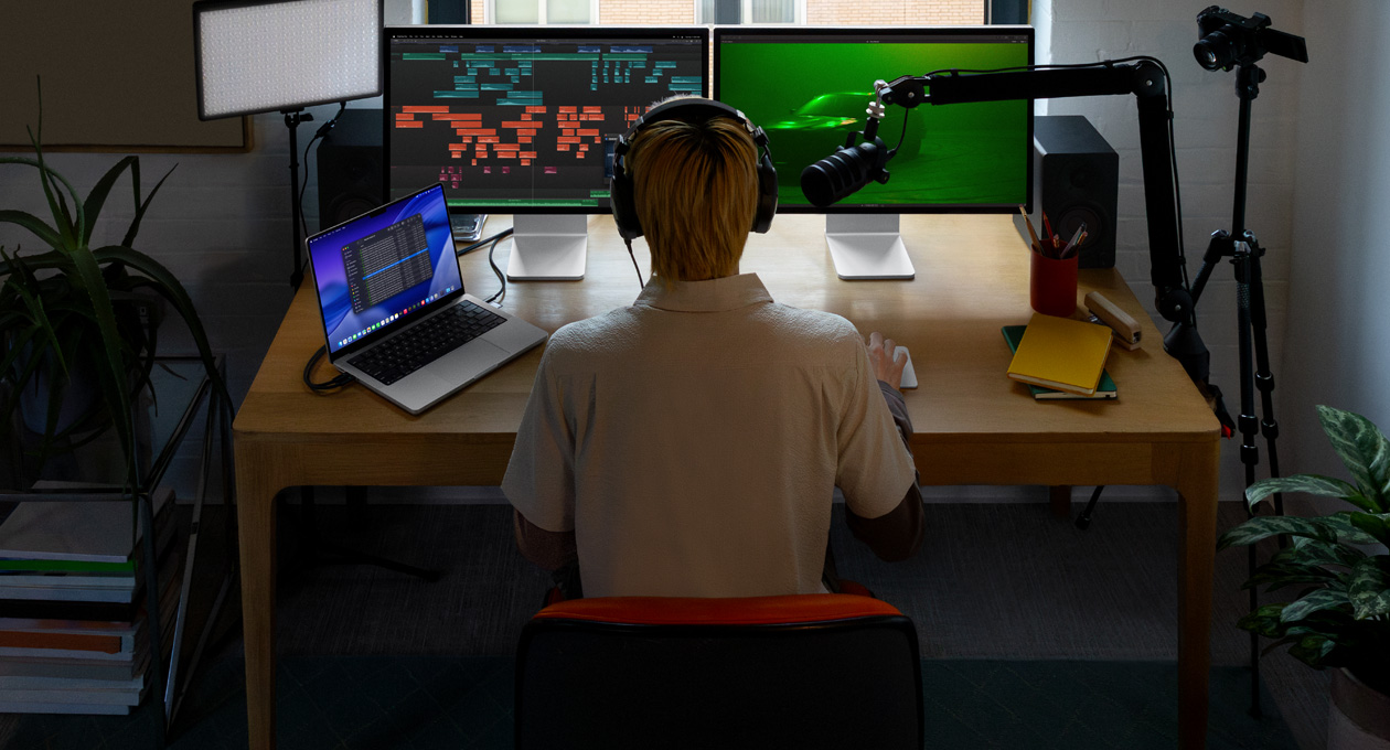 Person sitting at desk with a mic and headphones in front of three devices, Mac Studio Display screen, audio editing tools, sound clips in different blocks of color, a second Mac Studio Display screen showing, moody shot of car in fog with green lighting, MacBook Pro 16 screen showing Finder app on desktop showing Files for Final Cut Pro, dock, blue stylized desktop background
